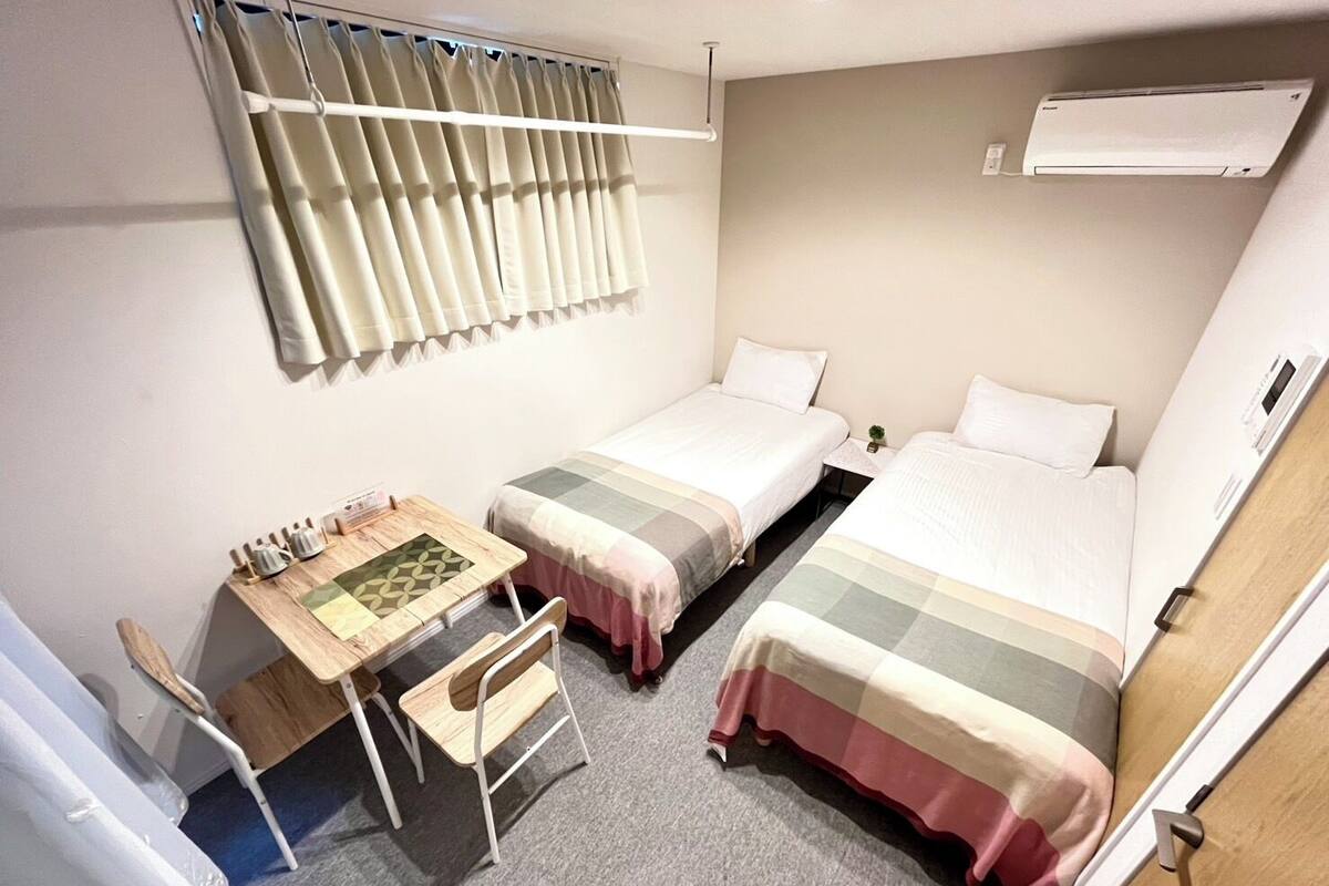 A small, compact room with two single beds, a small desk with a chair, and a window with curtains.