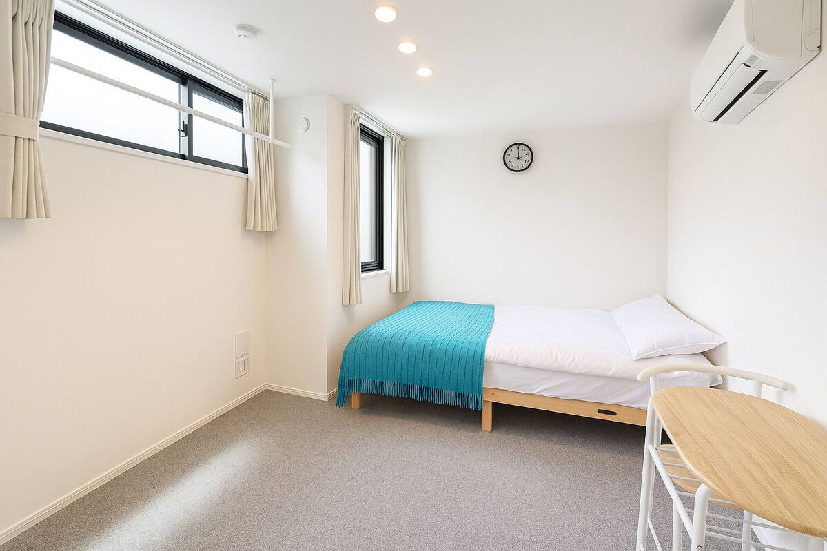 A small, neatly arranged bedroom with a single bed, a bedside table, and a chair. There is a window with curtains, a wall clock, and an air conditioning unit.