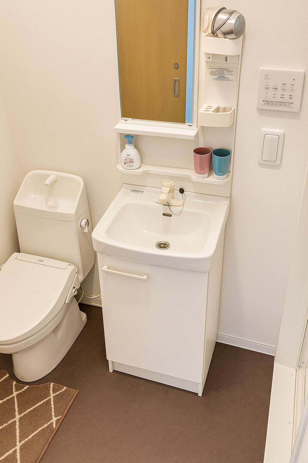 A small, white bathroom with a toilet, sink, and mirror. There are two cups on the sink's shelf, a soap dispenser, and a wall-mounted light fixture.