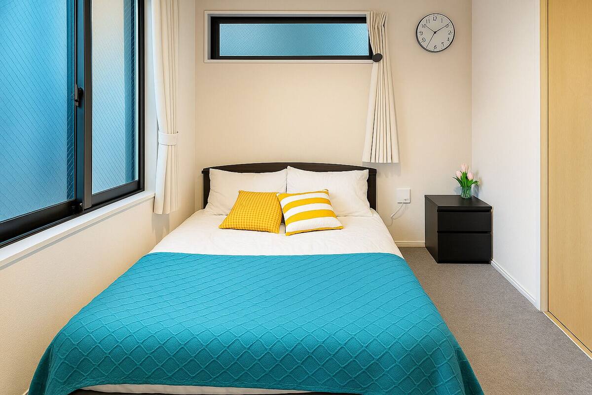 A neatly arranged bedroom with a bed, a bedside table with a lamp, a clock, and a window with curtains.