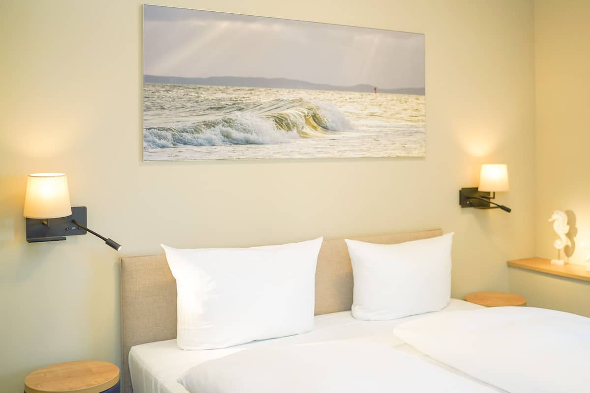 Camera d'albergo con un letto, due cuscini bianchi, una testiera, un quadro appeso raffigurante una spiaggia, due lampade a parete e un piccolo ripiano con un oggetto decorativo.