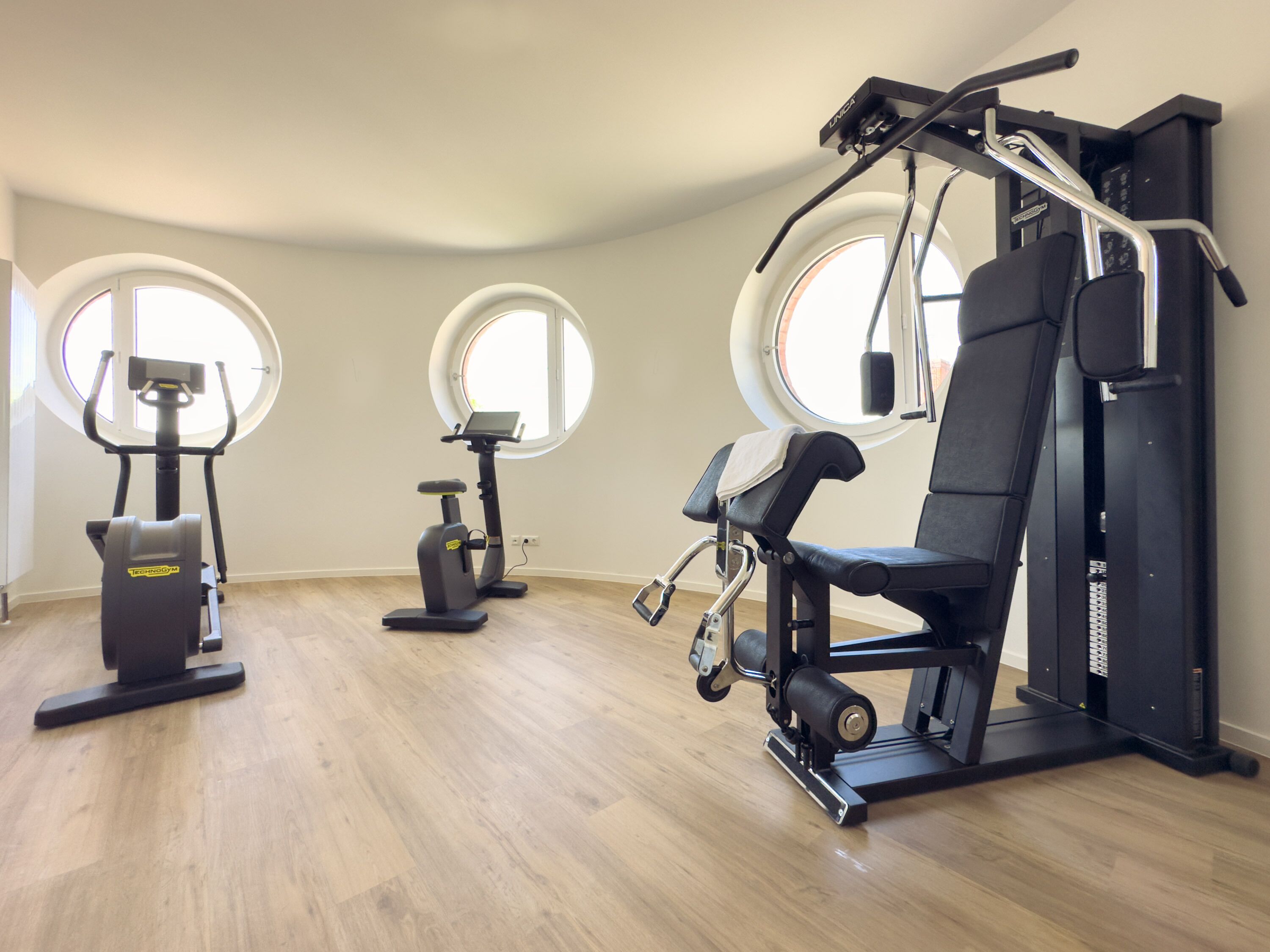 A gym with exercise equipment, large windows, and a wooden floor.