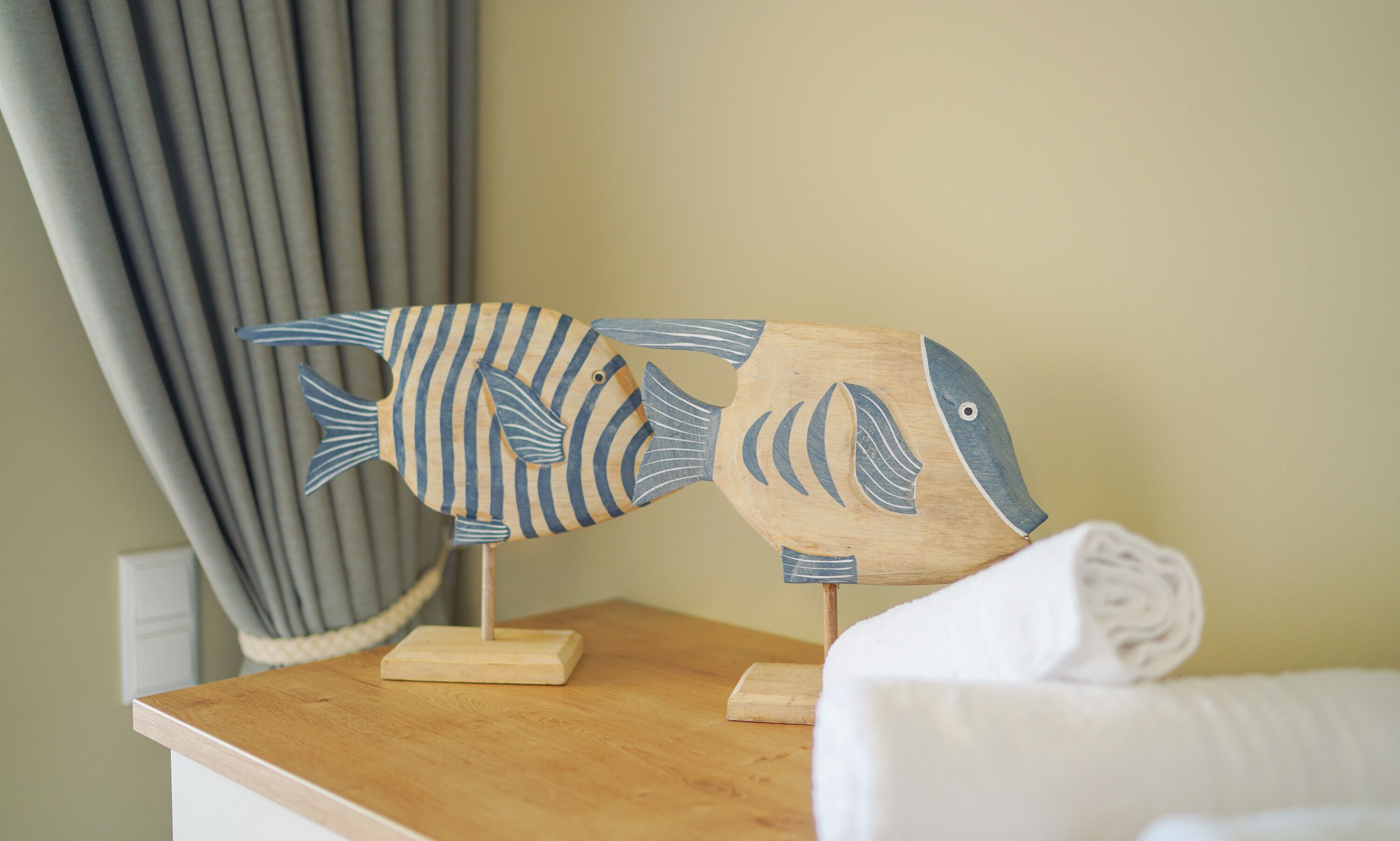 A wooden fish-shaped decoration on a wooden table in a room with a curtain and a white pillow.