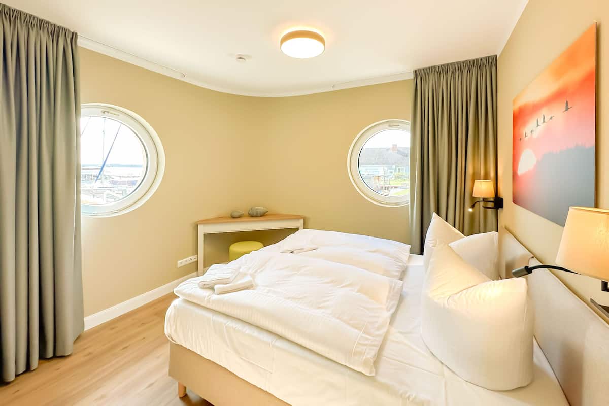 Una camera d'albergo con un letto, due finestre, un comodino, una lampada e un quadro appeso al muro.