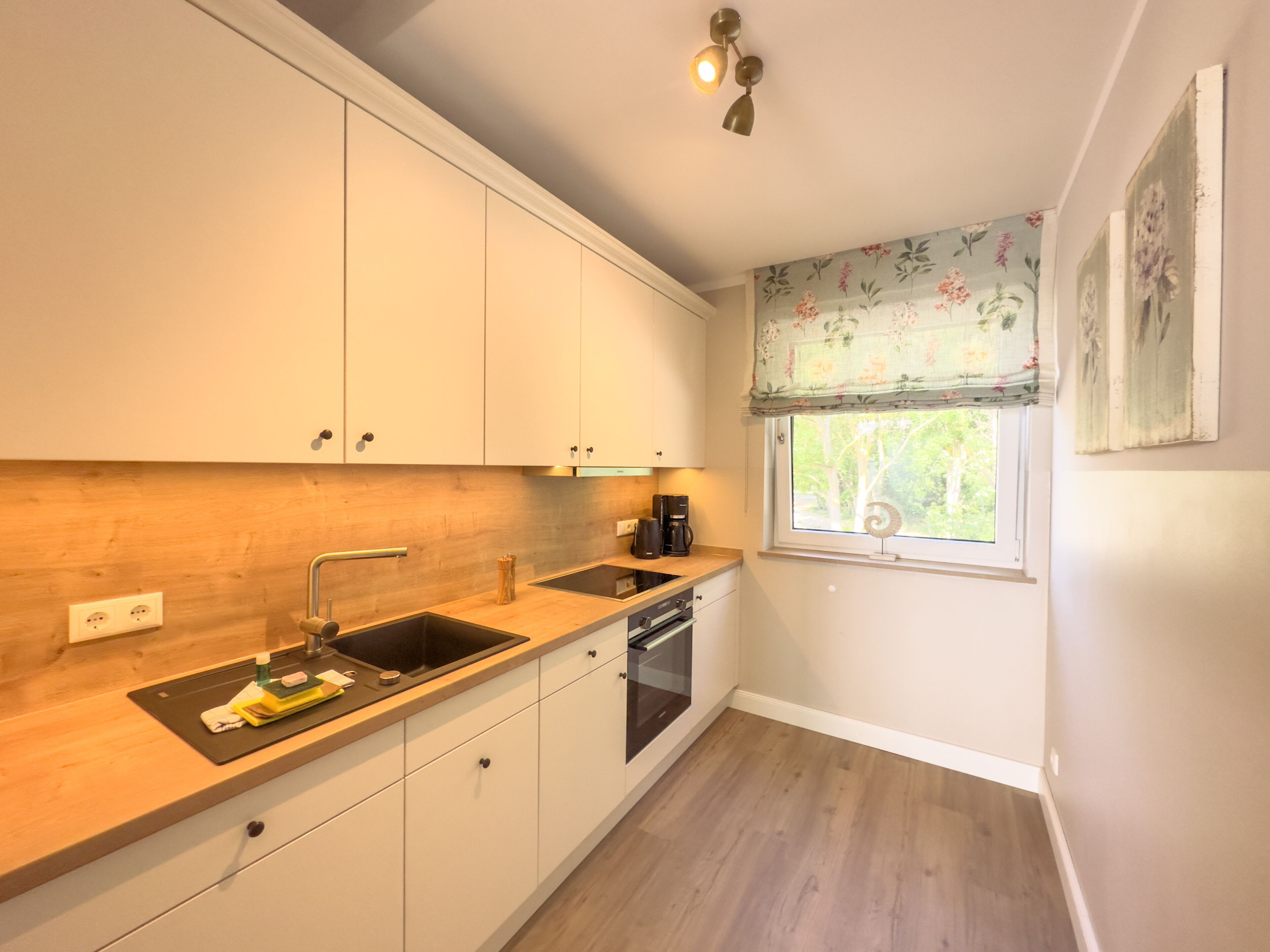 Une cuisine compacte avec des meubles blancs, un plan de travail en bois, un four encastré et une fenêtre agrémentée de rideaux à motifs floraux.