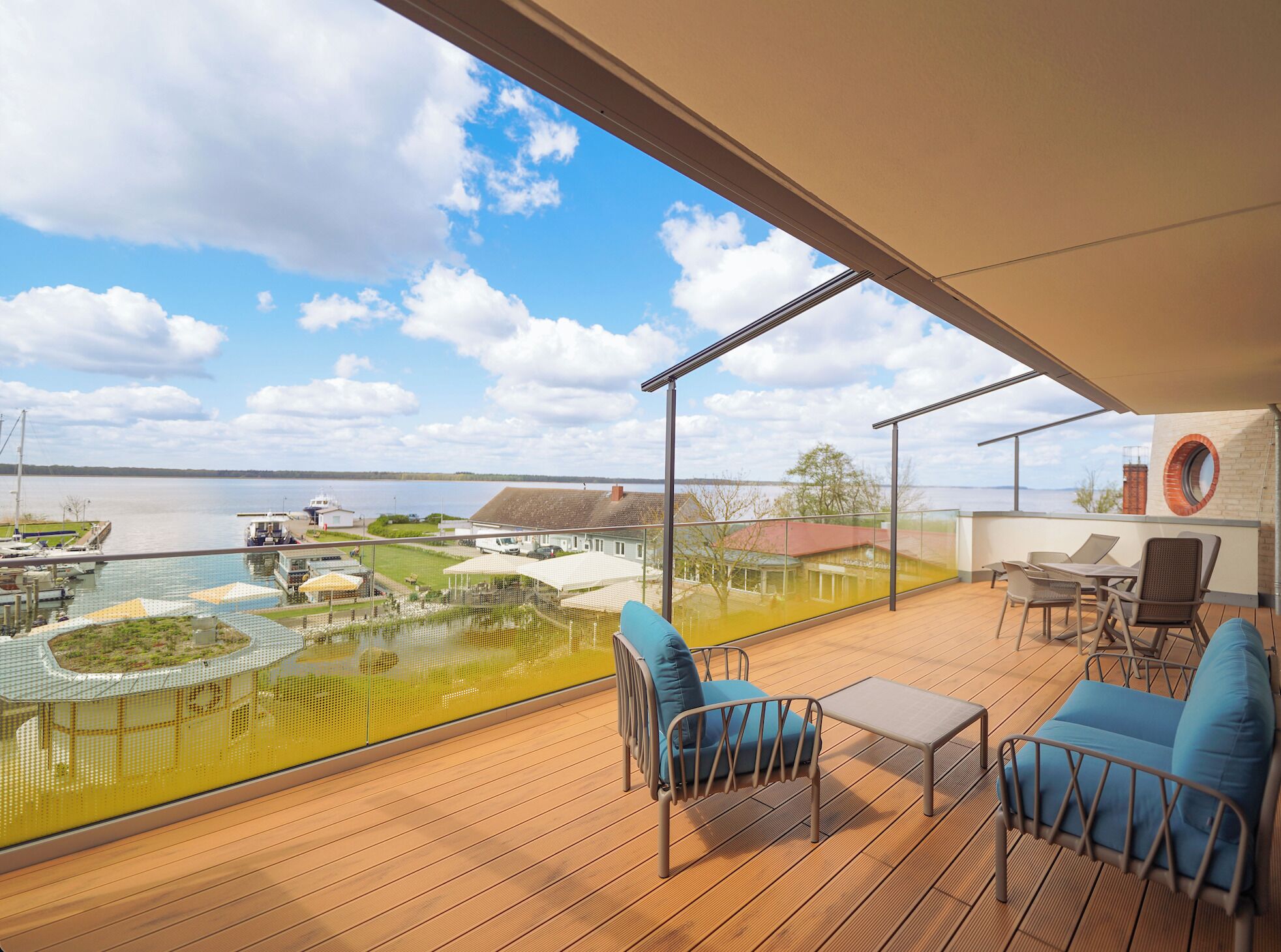 Un balcon spacieux doté d’un plancher en bois, de chaises bleues avec des coussins et d’une table. On peut admirer une vue dégagée sur une marina et les bâtiments situés de l’autre côté de l’eau.