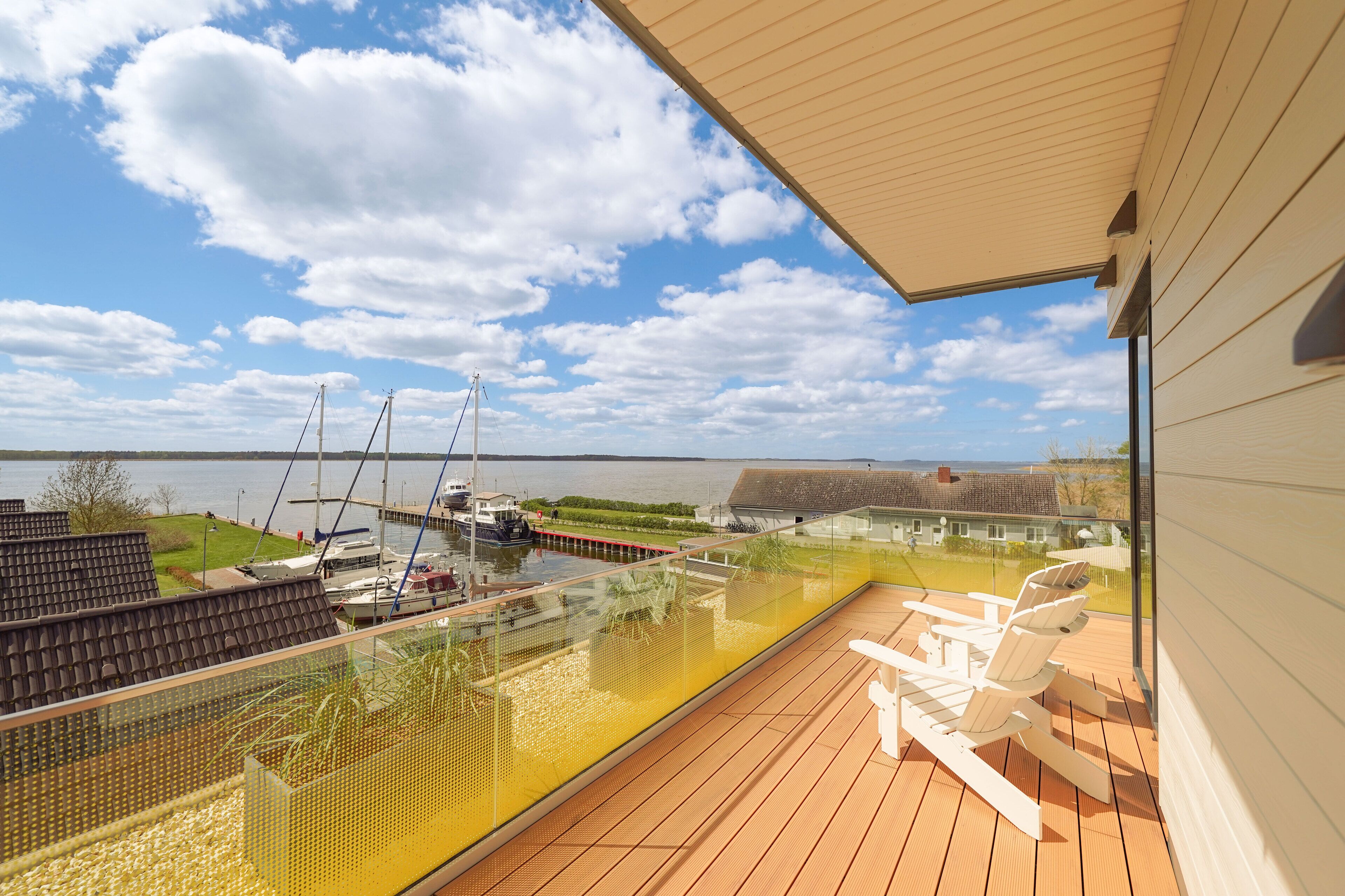 Una terraza de madera con barandilla de vidrio que da a un puerto deportivo con barcos y un cielo despejado.