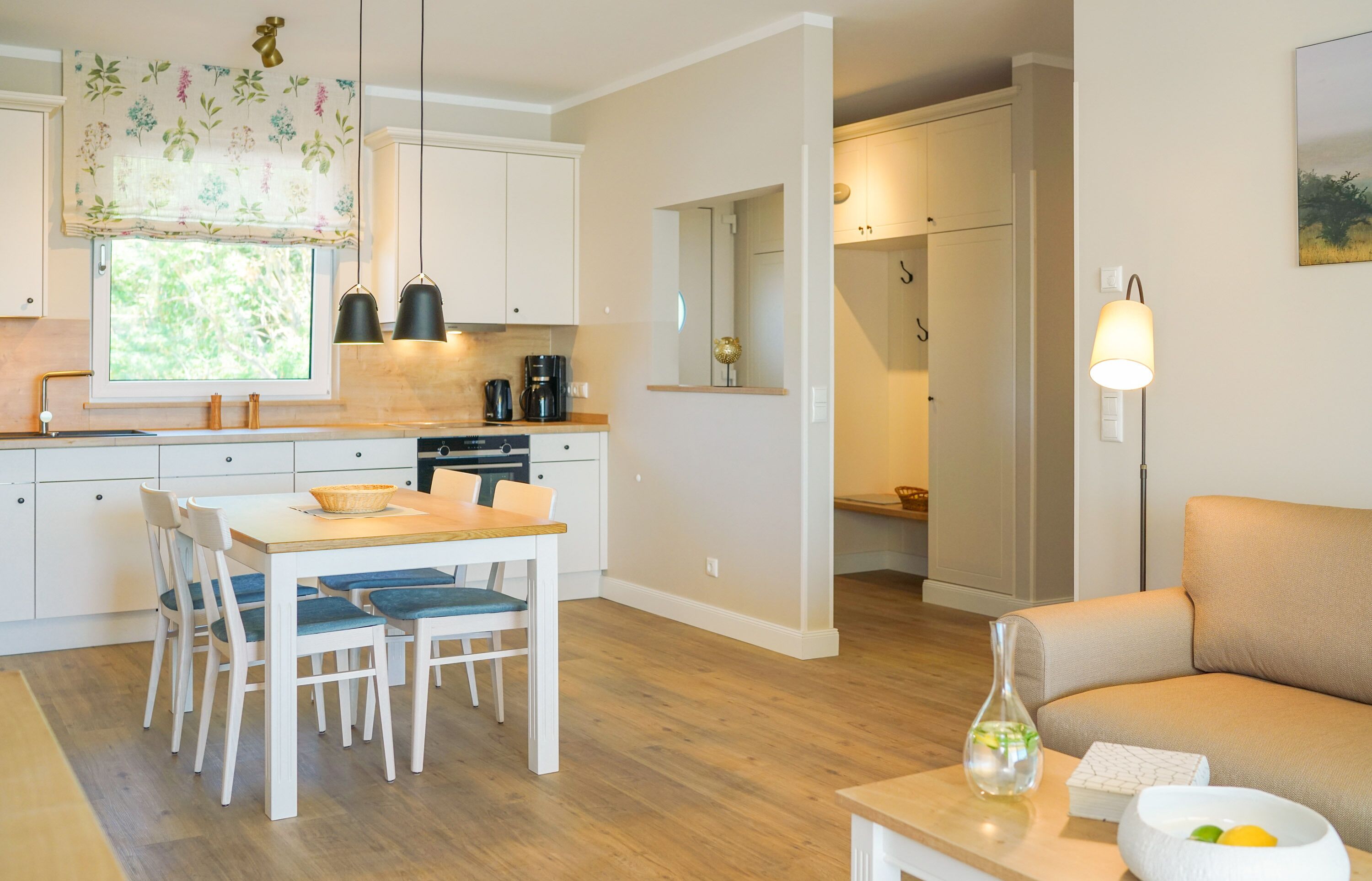 Een moderne keuken met witte kastjes, een eettafel met stoelen, een koffieapparaat en een raam met een bloemen gordijn.