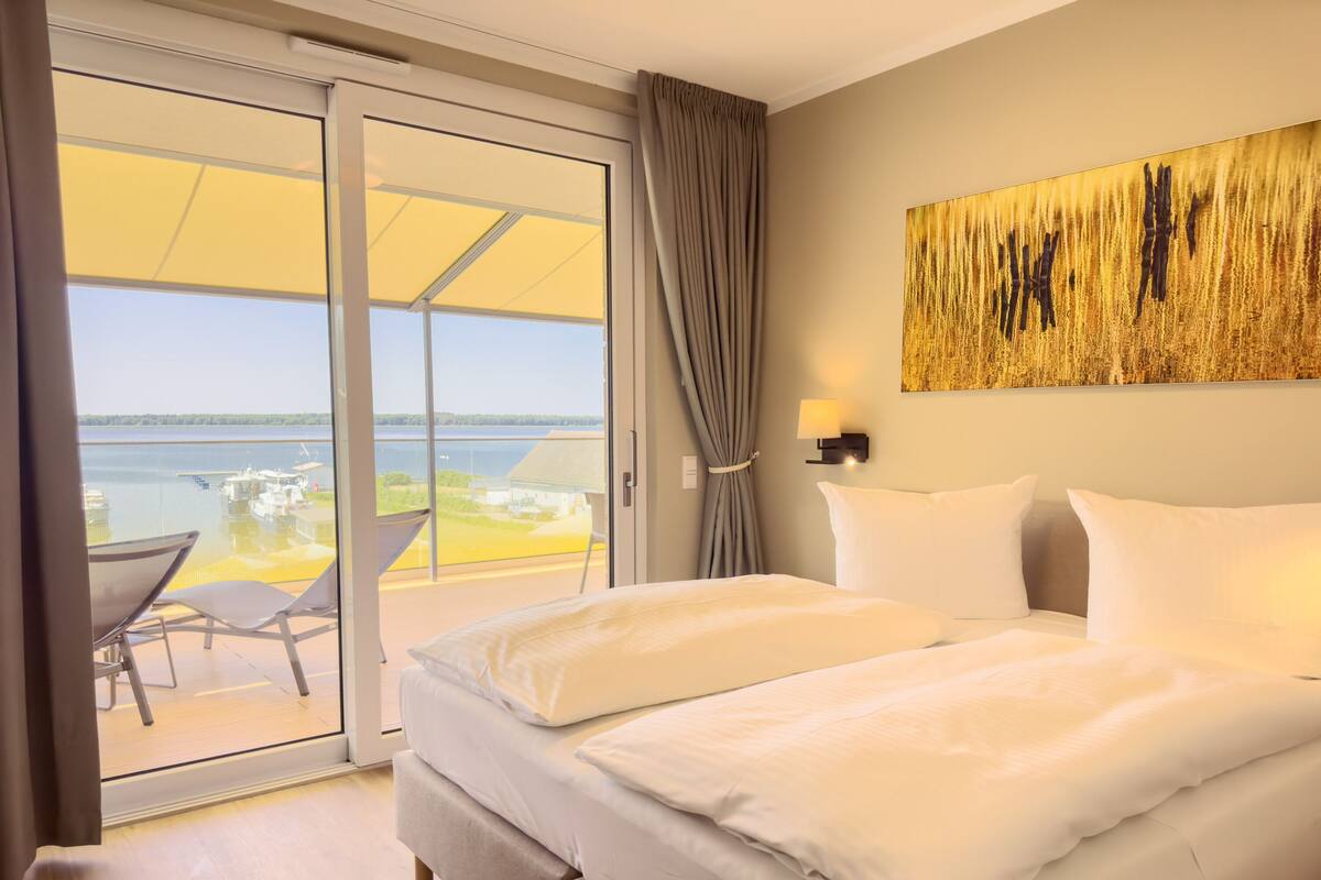 Una camera d'hotel con un letto, vista sul lago, zona pranzo con tavolo e sedie, e un grande quadro appeso al muro.