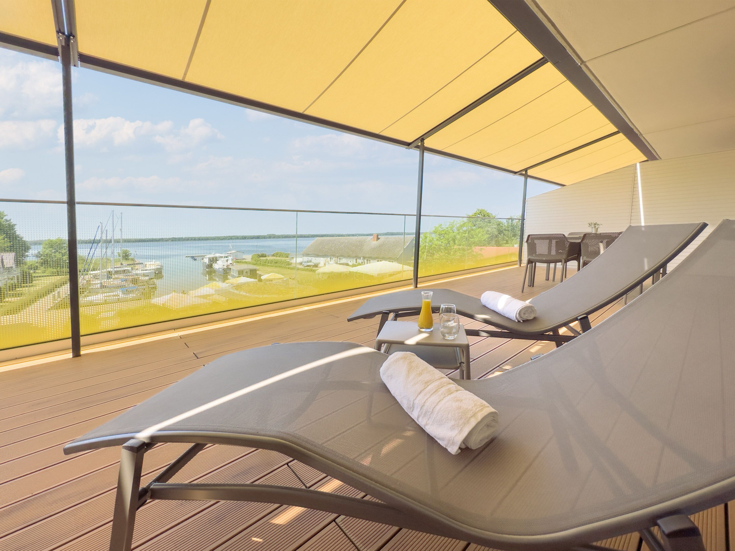 A balcony with two lounge chairs, a small table with a glass of juice and a bottle, and a view of the water and buildings.