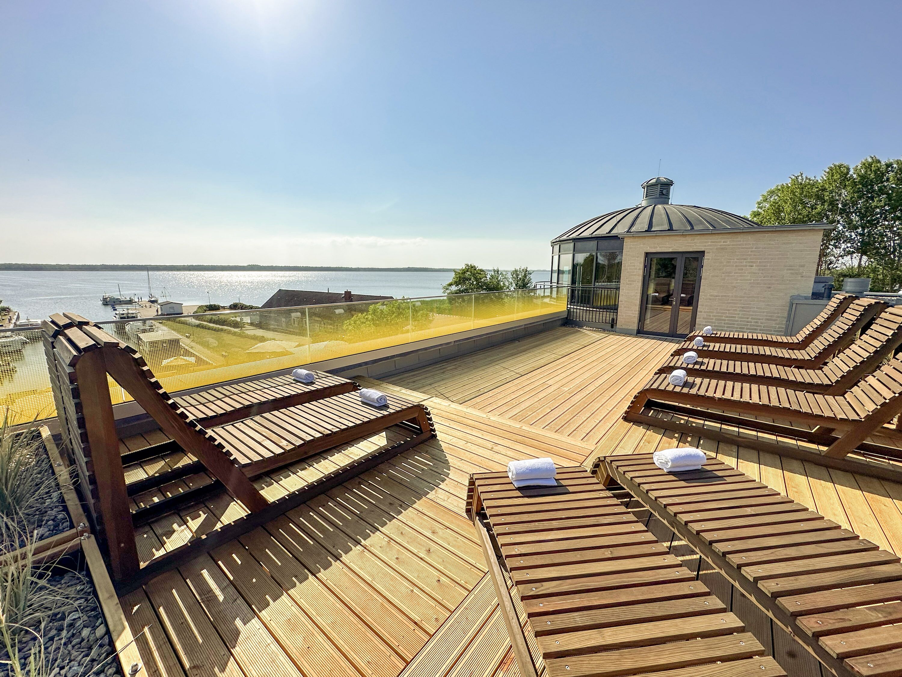 Eine Holzterrasse mit Liegestühlen und einer Glasveranda mit Blick auf ein Gewässer.