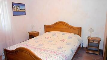 Un dormitorio con cama de madera, colcha estampada, mesitas de noche con lámparas, un cuadro en la pared y suelo de baldosas.
