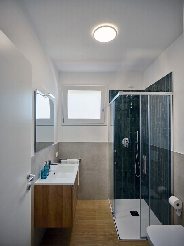A modern bathroom with a glass shower enclosure, a wooden vanity, a large mirror, and a window above the sink.