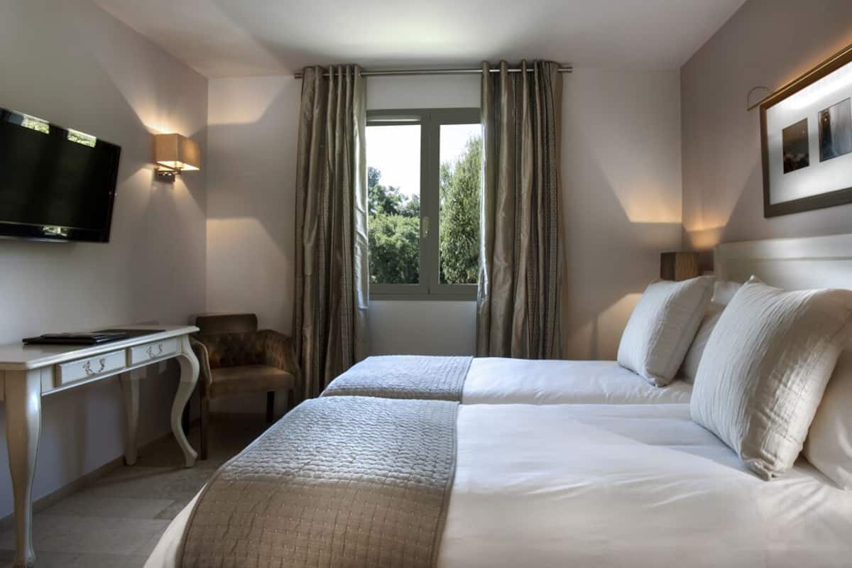 Ein Hotelzimmer mit zwei Betten, einem Schreibtisch, einem Stuhl, einem Fernseher und einem Fenster mit Vorhängen.