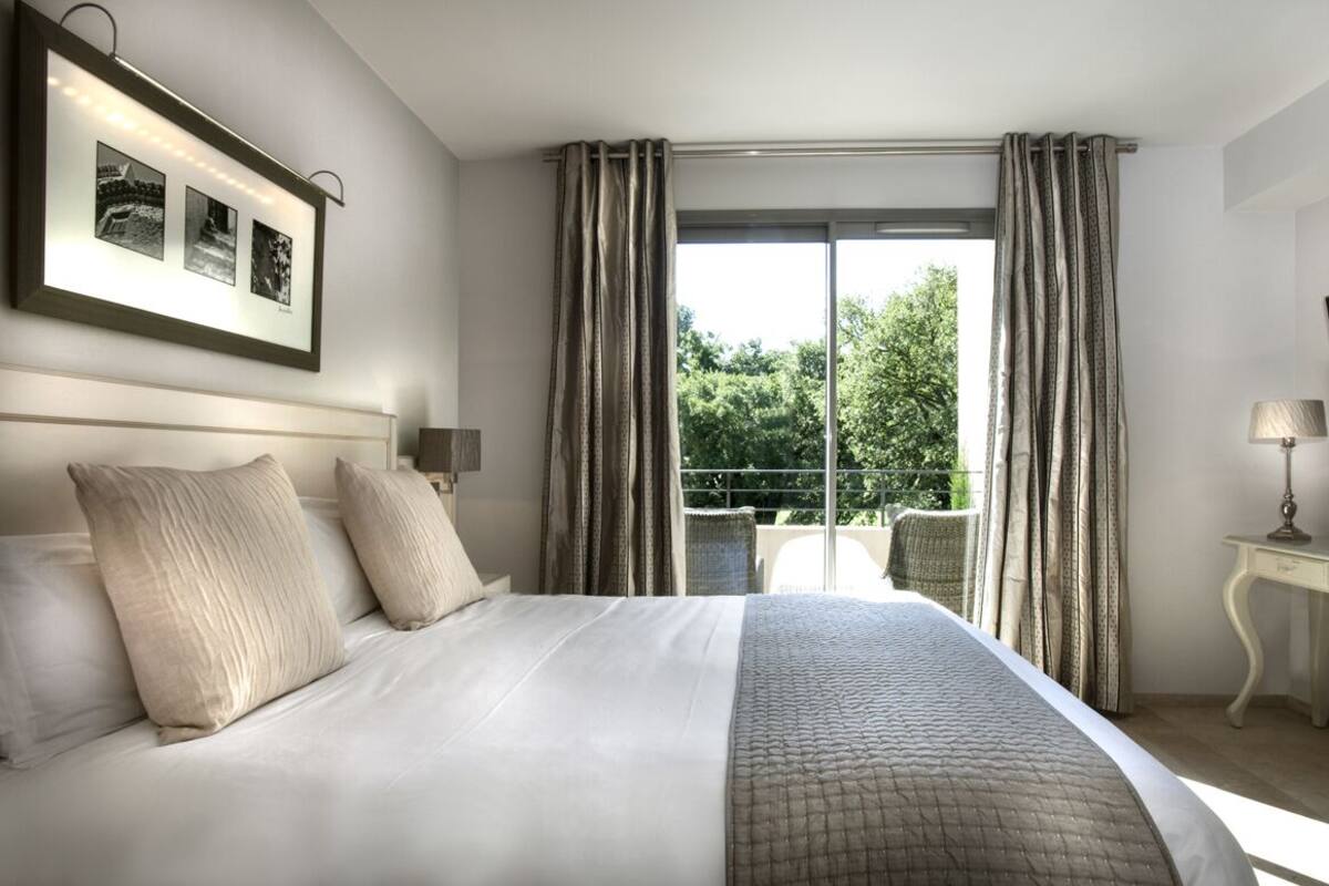 Ein Hotelzimmer mit einem großen Bett, einem Schreibtisch, einem Stuhl, einem Fernseher und einem Balkon mit Vorhängen.