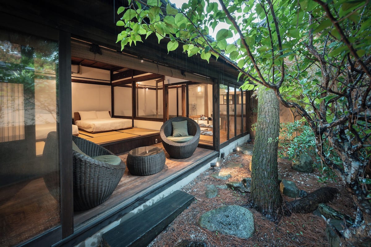 A traditional Japanese-style room with tatami flooring, a low wooden bed, and a sliding glass door leading to a private outdoor area with a wooden deck and a large tree.