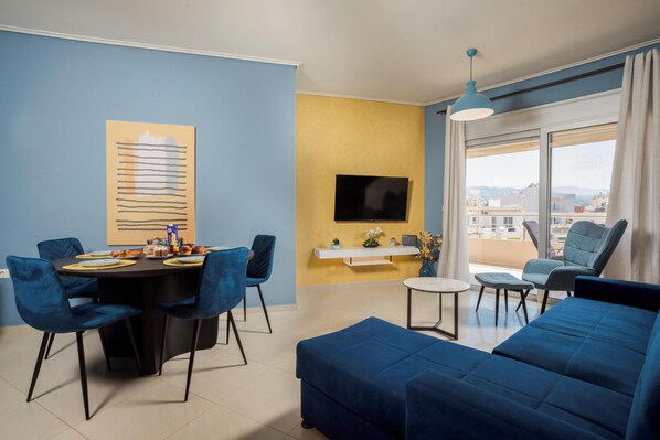A modern living room with a blue sofa, a dining table with chairs, a TV mounted on the wall, and a balcony with a view. - Zen City Beach Apartment (Chania)