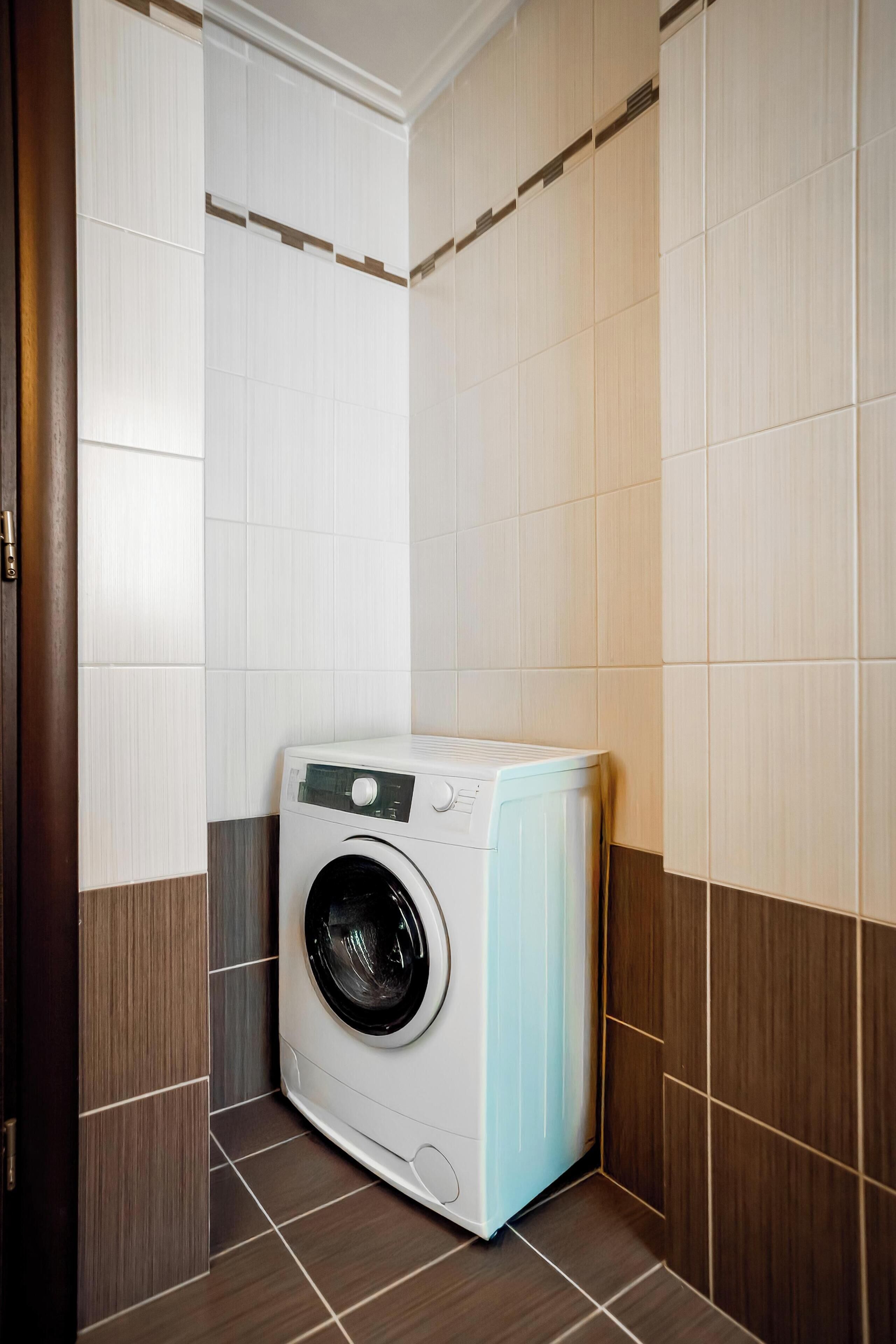 A compact laundry room with a front-loading washing machine, wooden panel walls, and dark brown tiled flooring.