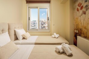A hotel room with two beds, a window with a view of buildings, and a radiator. - Zen City Beach Apartment (Chania)