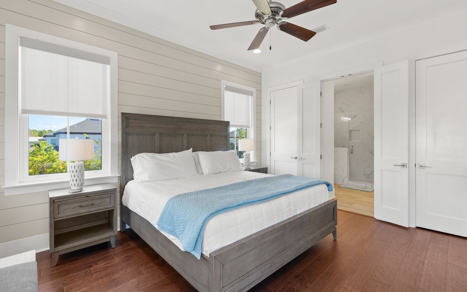 A bedroom with a bed, nightstands, a ceiling fan, and a view of the outside.