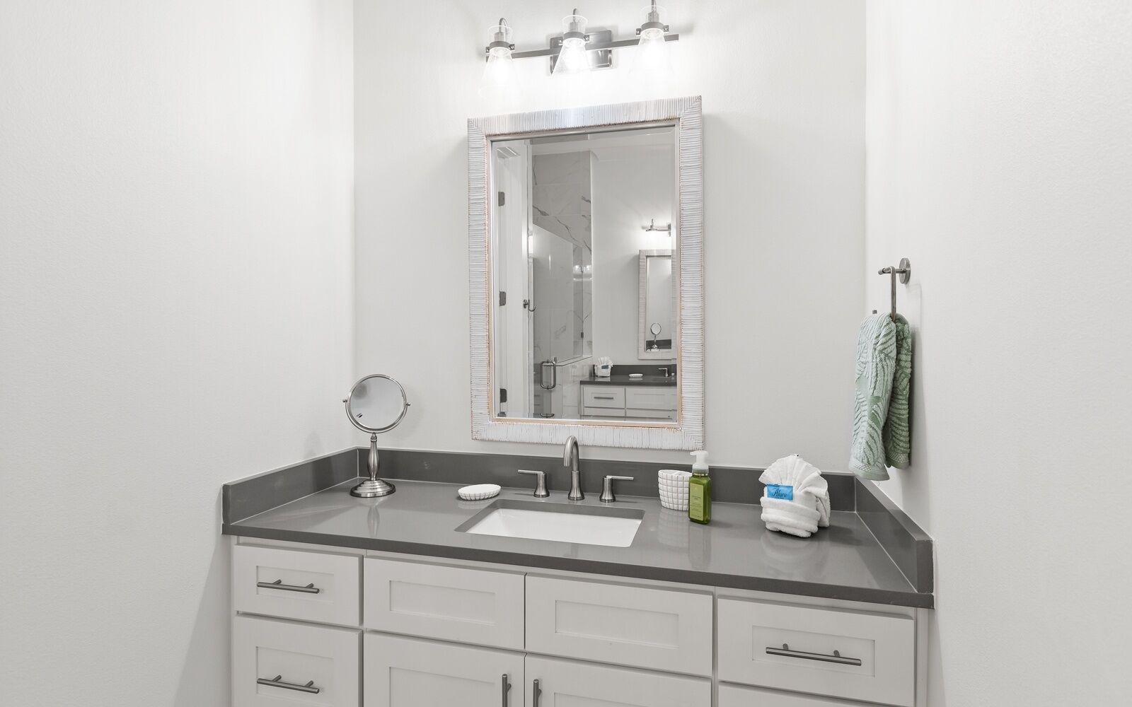 A modern bathroom with a large mirror, a round vanity mirror, a sink, and a towel rack with a green towel.