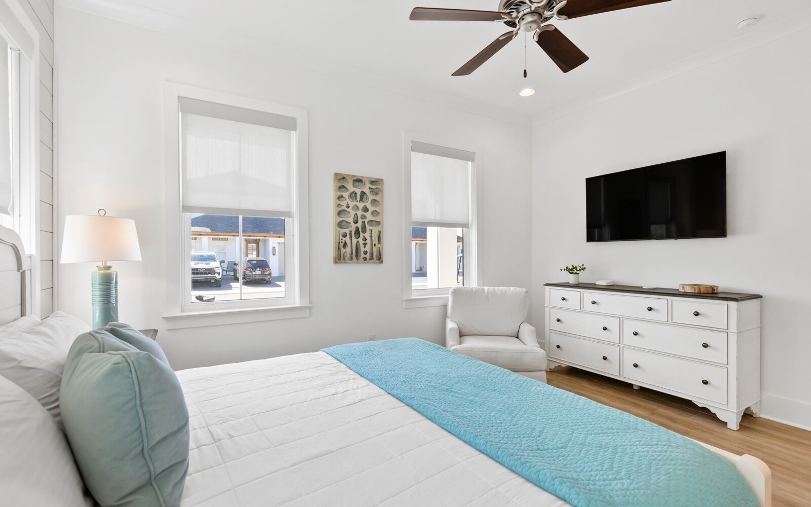 A bedroom with a bed, a dresser, a chair, a ceiling fan, and a TV on the wall.