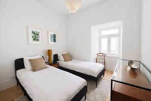 Ein Zimmer mit zwei Betten, einem kleinen Tisch, einer Lampe und einem Fenster.