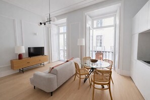 Ein modernes Wohnzimmer mit einer Couch, einem Esstisch, Stühlen, einem Fernseher auf einem Holzschrank und großen Fenstern.