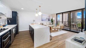 Une cuisine moderne dotée d’un îlot central, un coin repas avec un canapé et des fauteuils, et un balcon offrant une vue sur les immeubles.