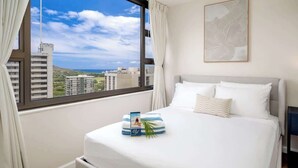 Une chambre d’hôtel dotée d’une grande fenêtre, d’un lit avec du linge de maison blanc, d’une œuvre d’art encadrée au mur et d’une petite table sur laquelle se trouvent un livre et une plante.