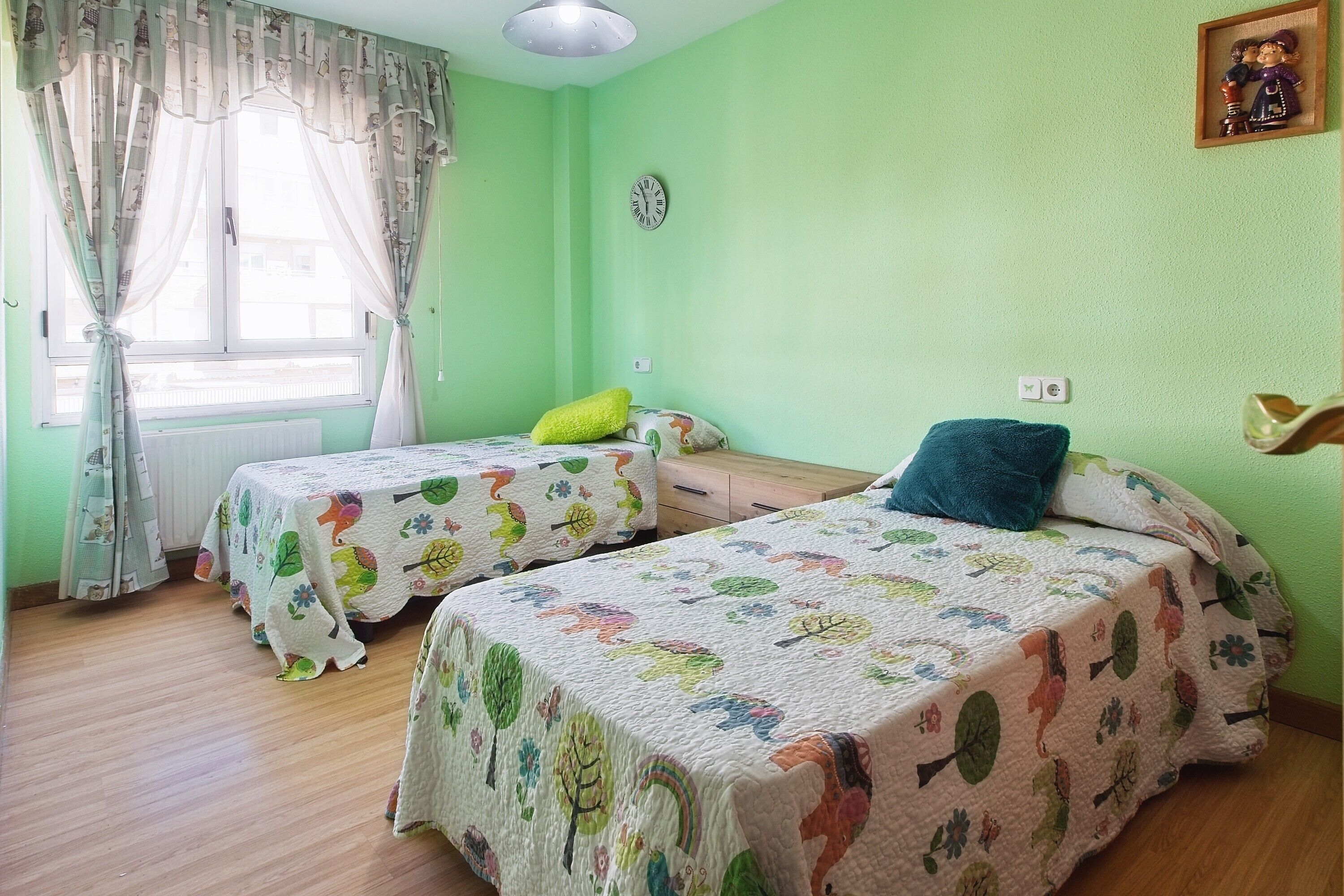 Una habitación con dos camas, una ventana con cortinas, un reloj y un cuadro pequeño en la pared.