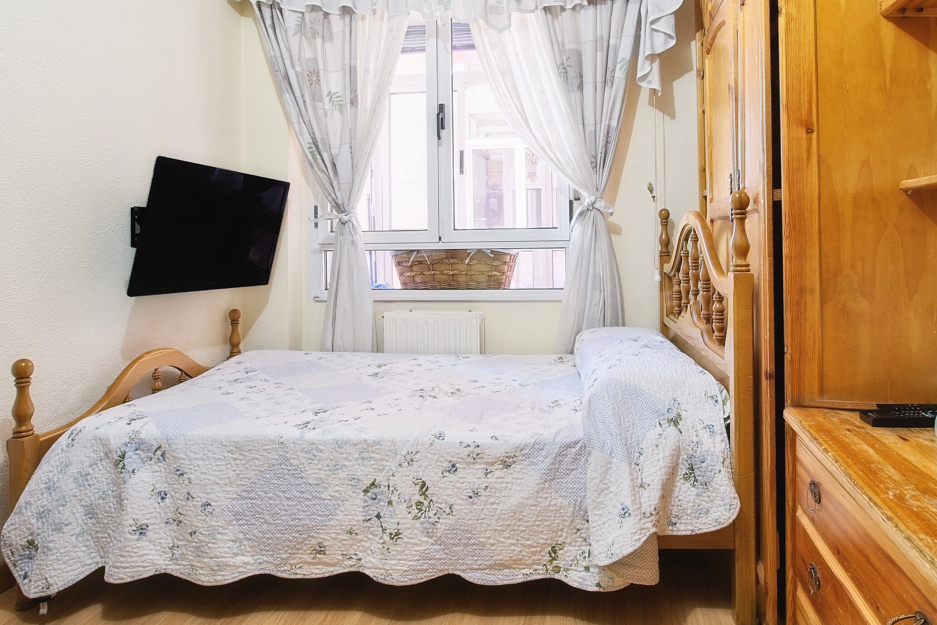 Un dormitorio con una cama, un televisor montado en la pared, una ventana con cortinas y un aparador de madera.