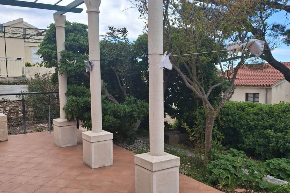 A covered outdoor area with concrete pillars, a tiled floor, and a garden with various plants.
