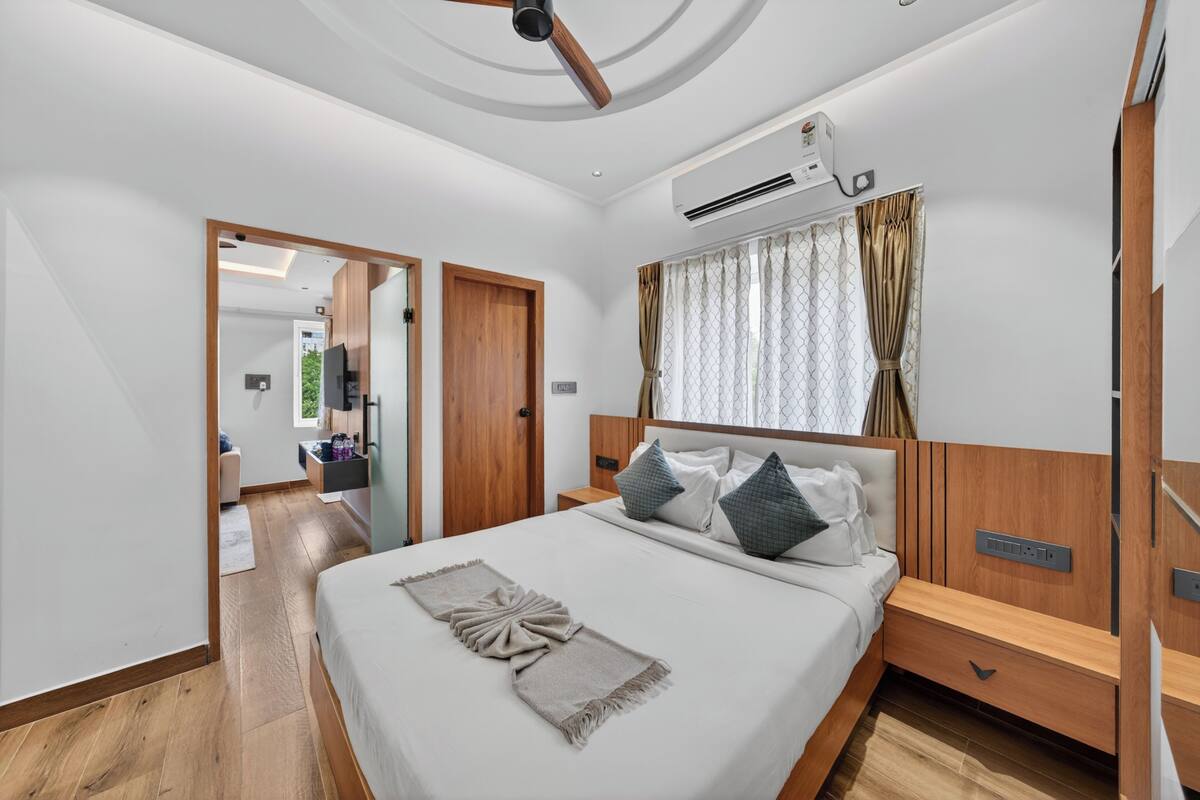 Quarto de hotel moderno com uma cama grande, cabeceira de madeira, mesas de cabeceira, ventoinha de teto e ar condicionado.
