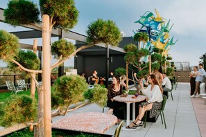 An outdoor seating area with round tables and chairs, surrounded by small trees and a decorative metal sculpture. - Steps to Mission Ballroom + Kitchen. Bar. Playroom (Denver)
