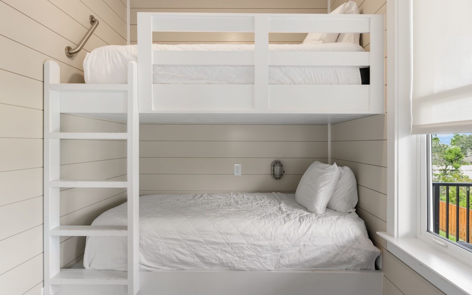 A bunk bed with a ladder, a bedside table, a wall-mounted lamp, and a window with a view of greenery.