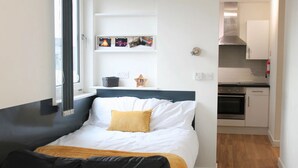 A bedroom with a bed, a bedside table, a window, and a view of a kitchen. - For Students Only Newarke St (Leicester)