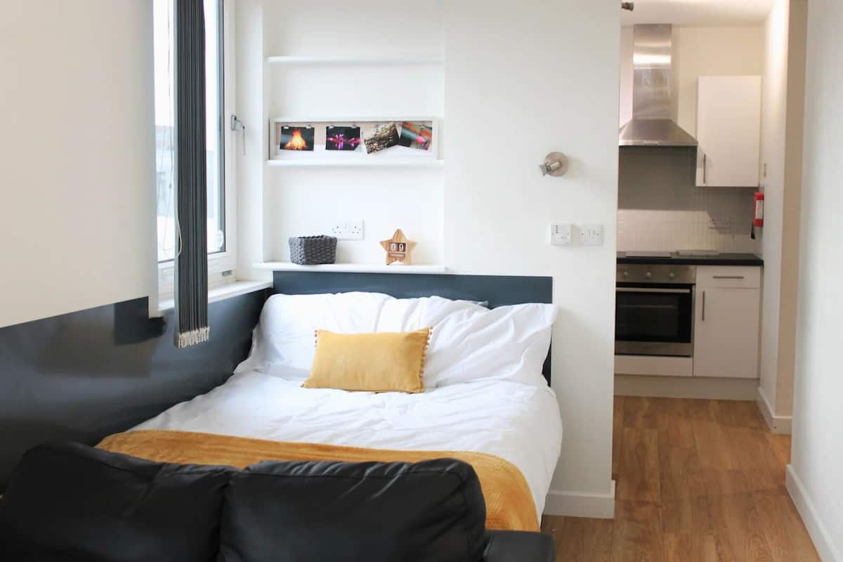 A bedroom with a bed, a bedside table, a window, and a view of a kitchen.