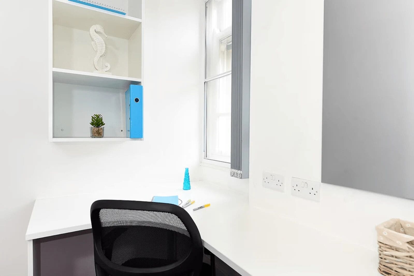 Un espacio de oficina moderno con un escritorio blanco, una silla de oficina negra, una planta pequeña en maceta y un caja de almacenamiento azul.
