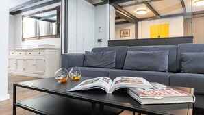 Uma sala de estar moderna com um sofá cinzento, uma mesa de centro com dois copos de líquido âmbar e uma revista aberta.