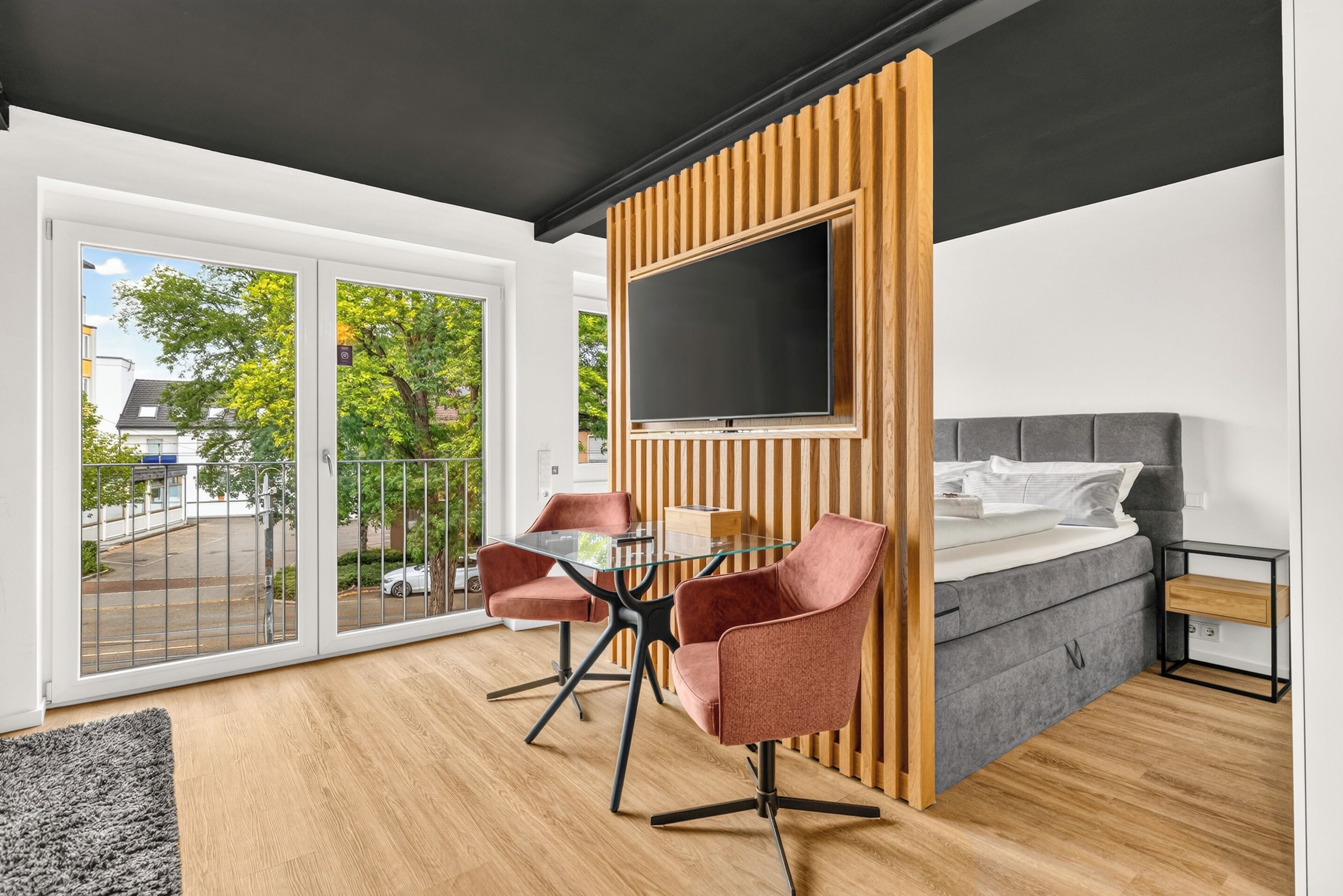 A modern hotel room with a bed, a TV, a dining area with chairs, and a view of the outside.