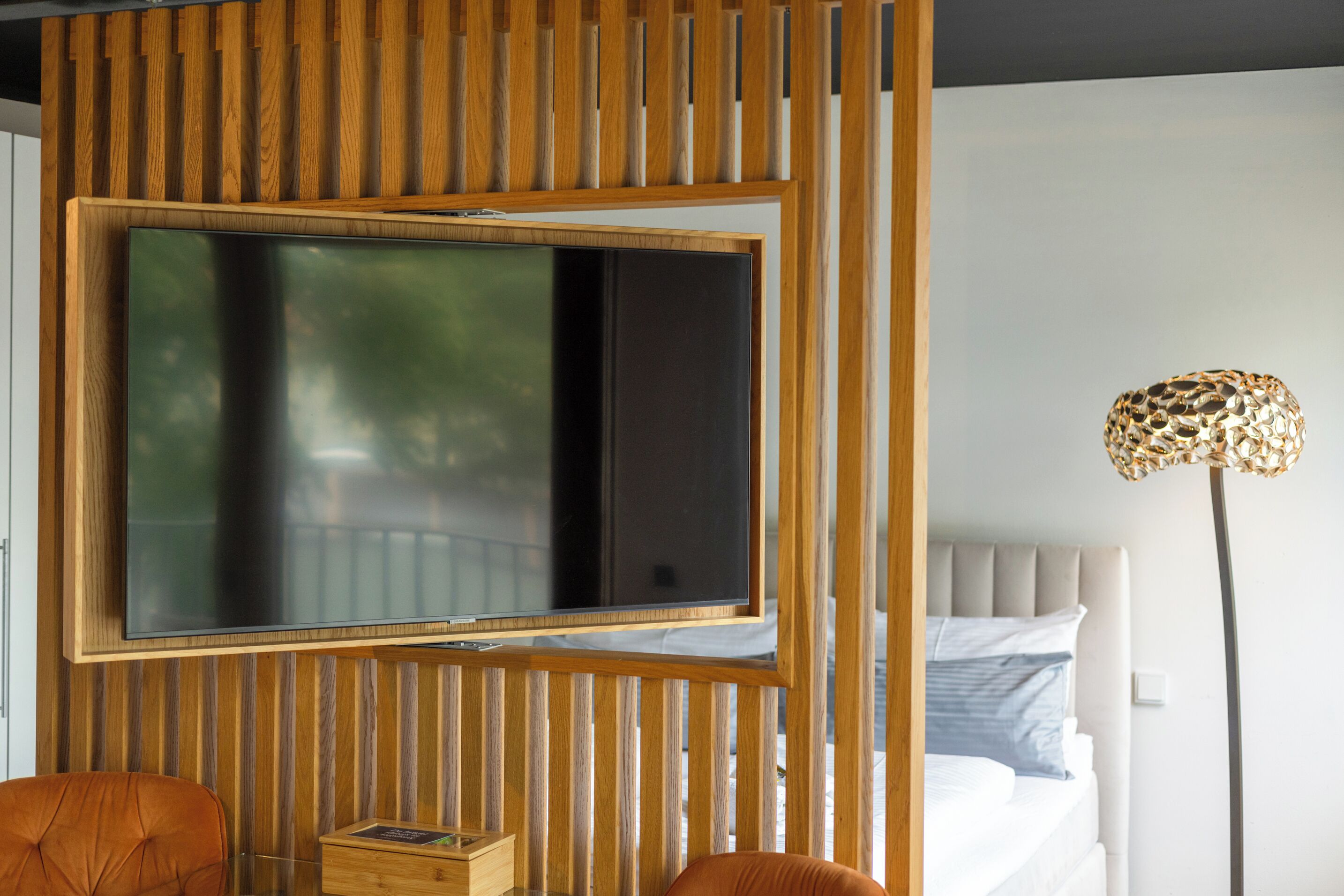 A room with a large flat-screen TV mounted on a wooden panel, a bed with a headboard, and a floor lamp.