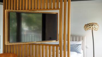 A room with a large flat-screen TV mounted on a wooden panel, a bed with a headboard, and a floor lamp.
