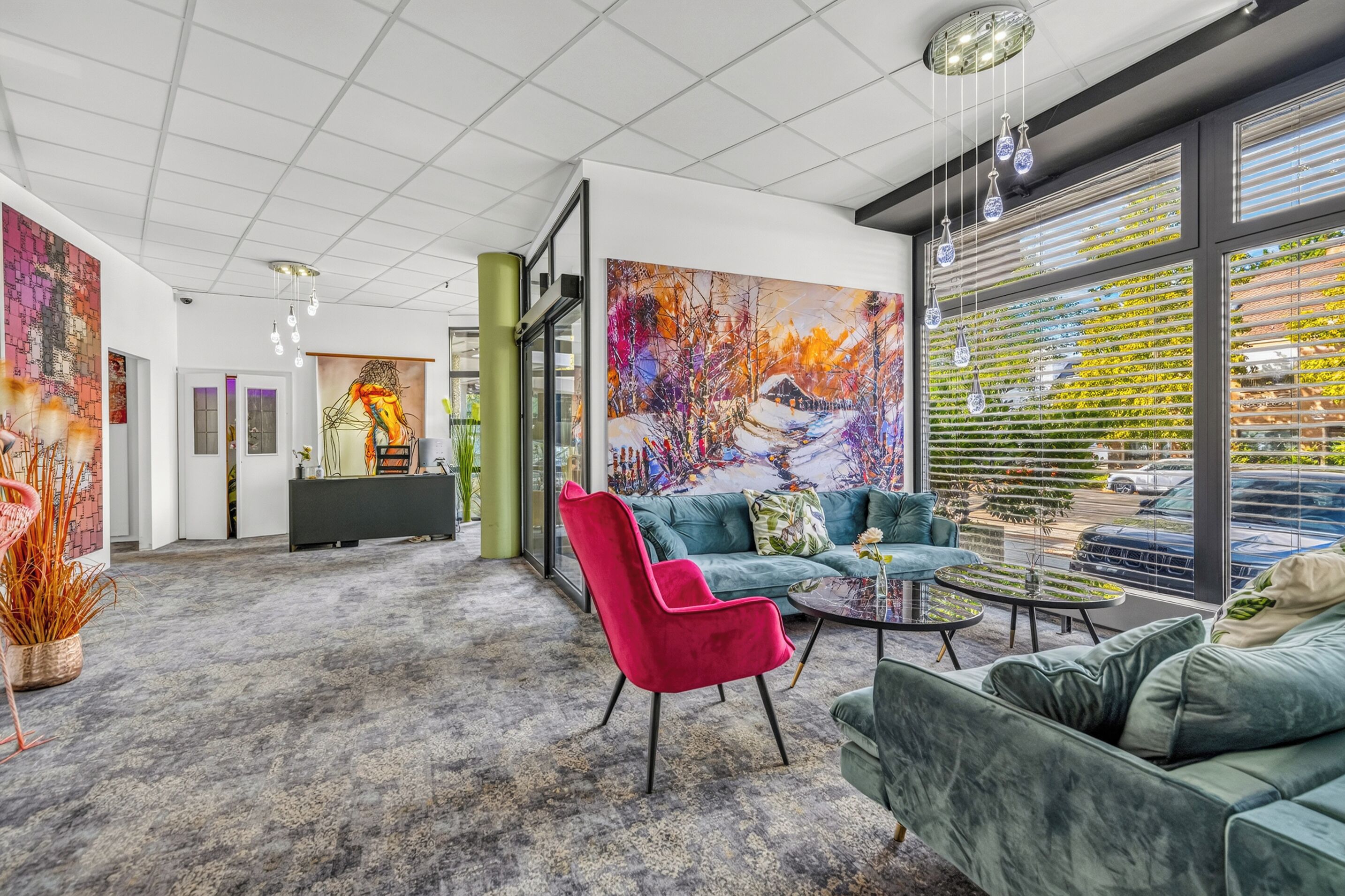 A modern interior with a grey carpet, a red chair, teal sofas, and large windows with blinds.