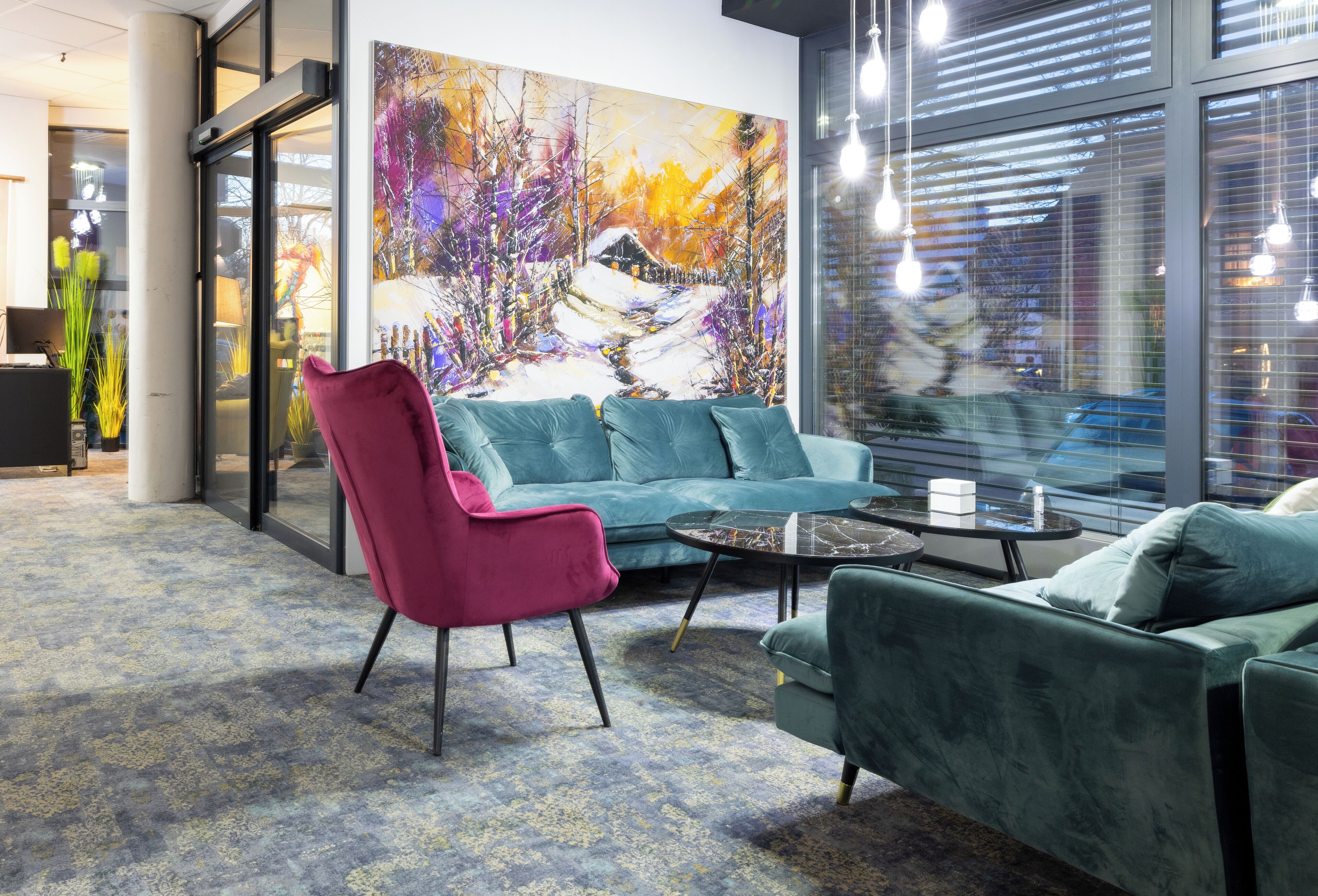 Un área de descanso moderna con un sofá azul, una butaca rosa y un sillón verde. Un cuadro abstracto de gran tamaño decora la pared, y hay una puerta de cristal que da al exterior.