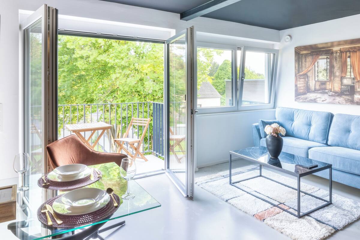 Ein modernes Wohnzimmer mit einem Glastisch, gedeckt für ein Essen, einem blauen Sofa und einem Balkon mit Gartenmöbeln.