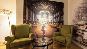 Une pièce avec deux fauteuils verts, une table en verre et un grand tableau représentant l’intérieur d’une bibliothèque.