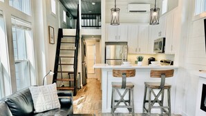 A modern kitchen with white cabinets, stainless steel appliances, and a central island with stools. A black leather sofa and a patterned rug are in the living area.
