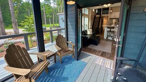 A wooden deck with Adirondack chairs, a blue patterned rug, and a view of a forested area.
