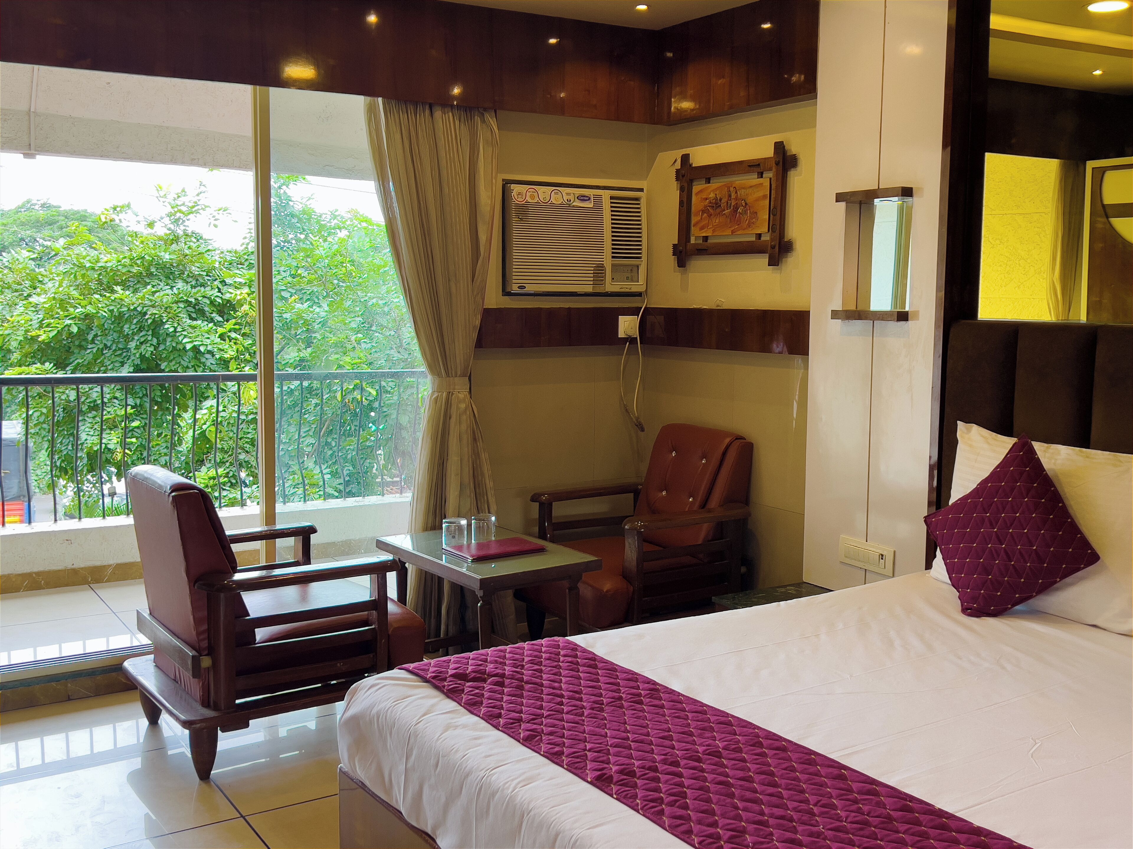 A hotel room with a bed, a desk, a chair, a window with a view of greenery, and a wall-mounted air conditioning unit.